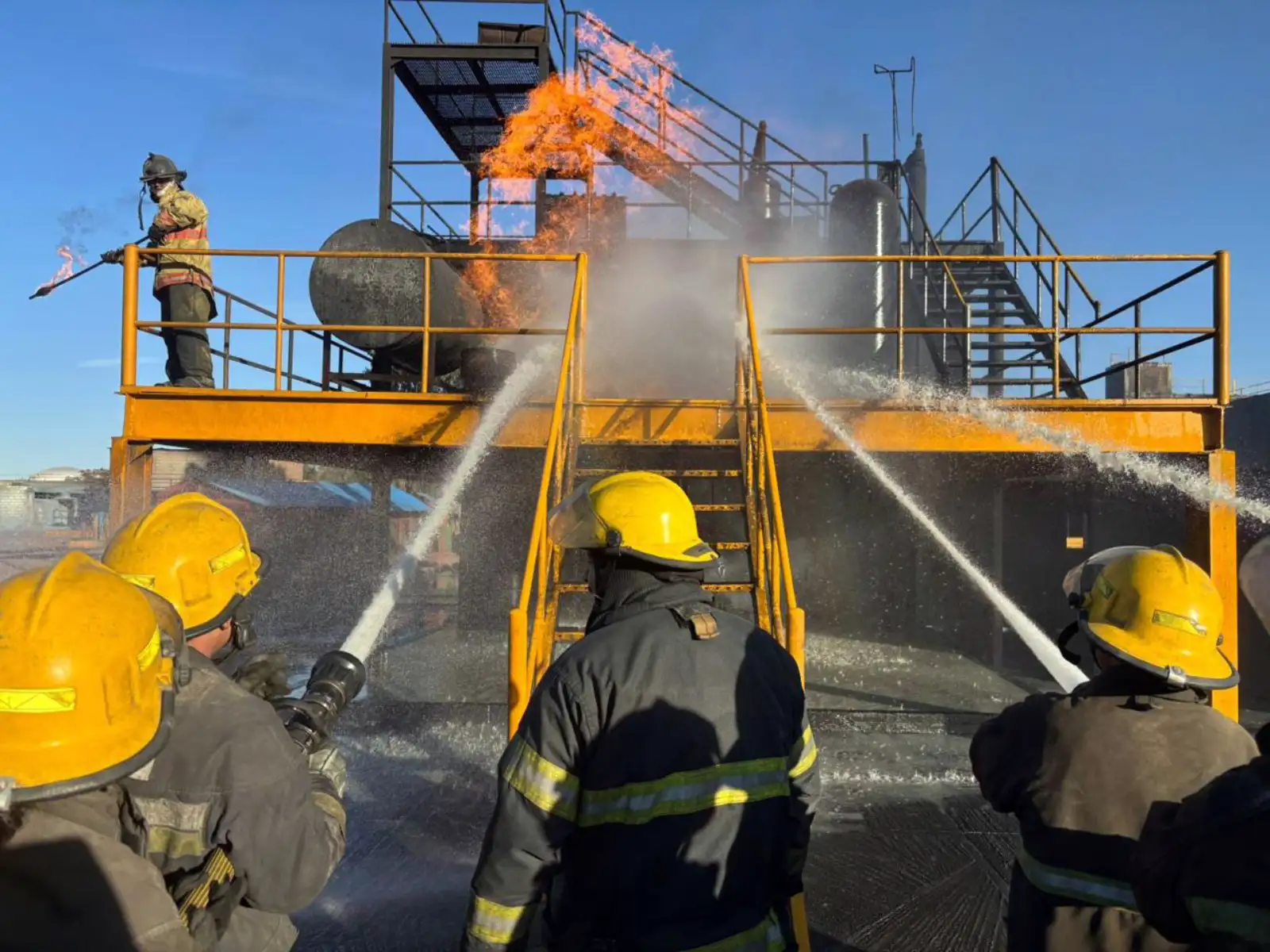 Brigadistas atacando incendio en torre de entrenamiento con líneas de agua